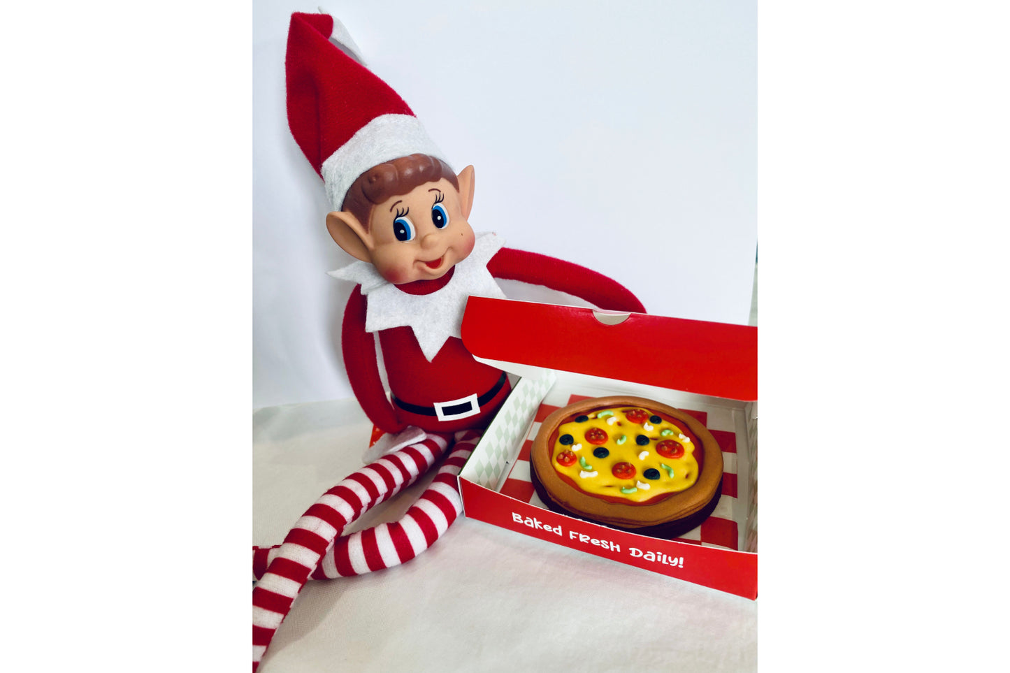 Elf on the shelf sat by open mini pizza box. Pizza made from biscuit and made to look like it has been delivered from the North Pole.
